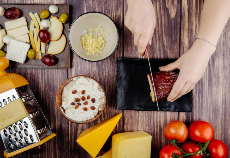 a woman cuts smoked cheese on a black tray cottage cheese in a bowl grater pickled olives and fresh tomatoes on rustic background top view